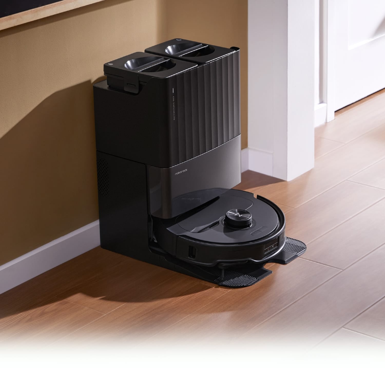Black Roborock Qrevo Master robot vacuum docked in its multifunctional auto-empty and mop washing station on a wooden floor.