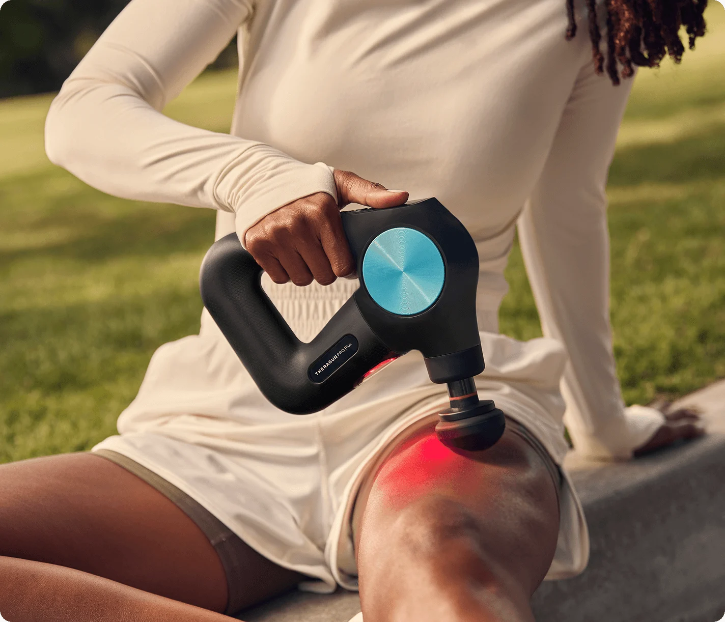 Woman using a black Theragun PRO Plus massager with a blue control disk and red light on her thigh outdoors.