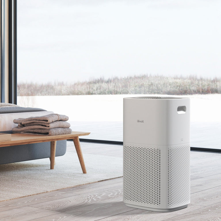 Modern white Levoit Core 600S smart air purifier in a clean bedroom setting with a large window overlooking a winter scene.