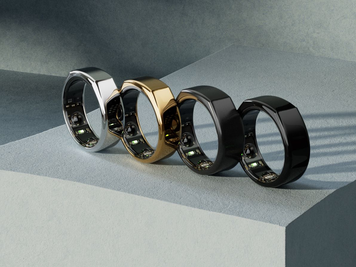 Four Oura Ring Gen3 smart rings: silver, gold, and two black, with visible internal sensors, on a textured gray surface.