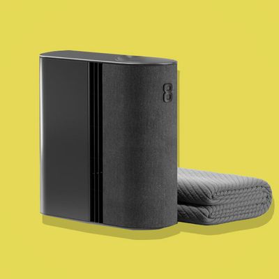 Eight Sleep Pod 4 smart sleep system's dark gray hub device beside a folded gray cooling mattress pad on a yellow background.