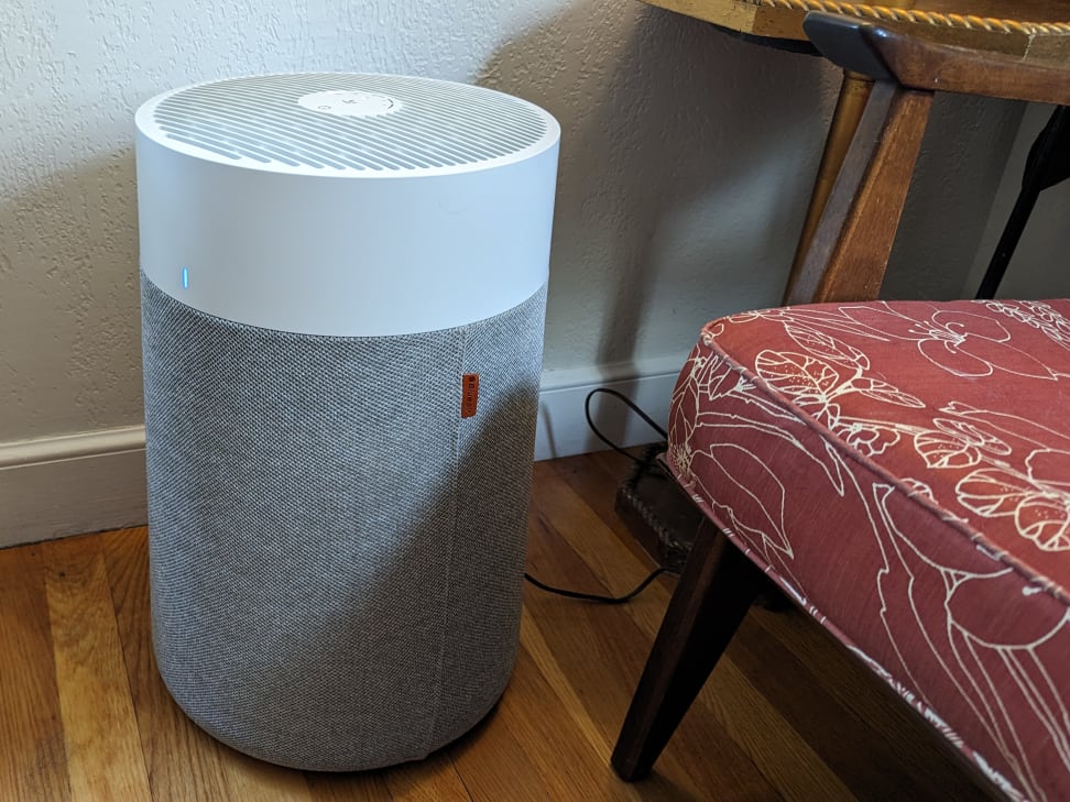 Blueair Blue Pure 311i Max air purifier with grey fabric pre-filter and white top, glowing blue indicator light, on a wooden floor.