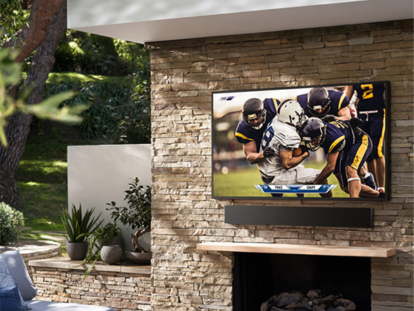 Outdoor patio features Samsung The Terrace TV mounted on a stone wall, playing a football game, with a soundbar.