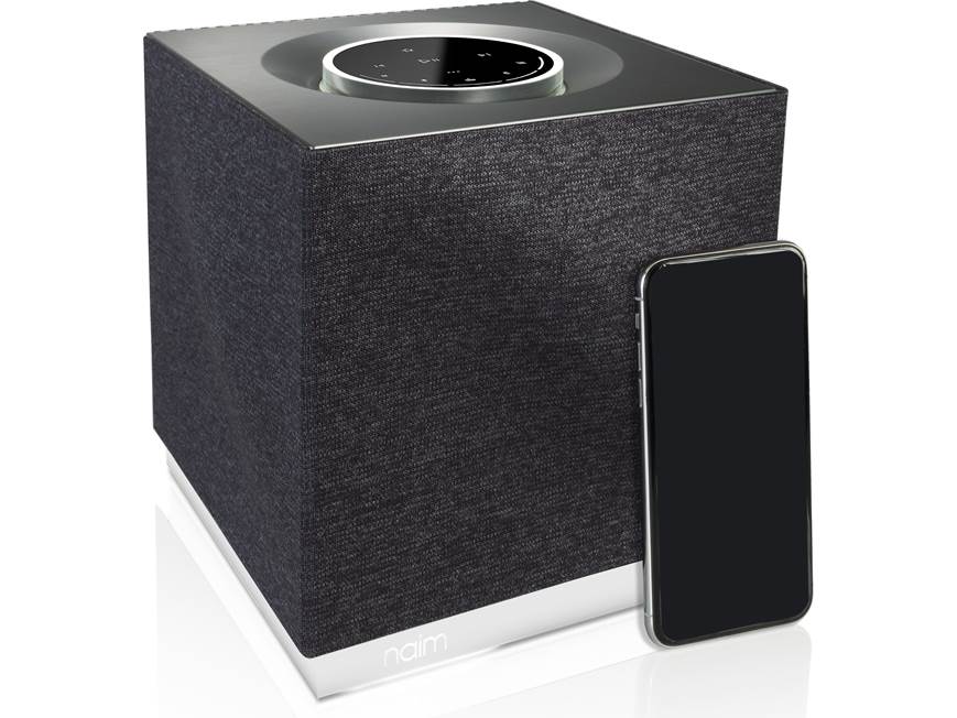 Dark gray Naim Mu-so Qb 2nd Generation wireless speaker with silver top control and a smartphone beside it.